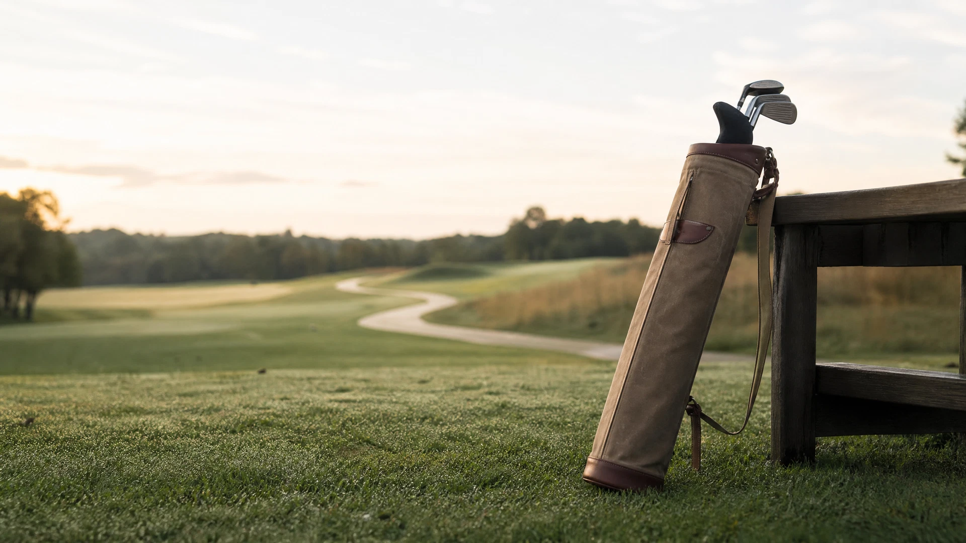 A premium minimalist Sunday pencil bag crafted from olive waxed canvas with white full-grain leather trim and brass hardware, leaning against a wooden bench.