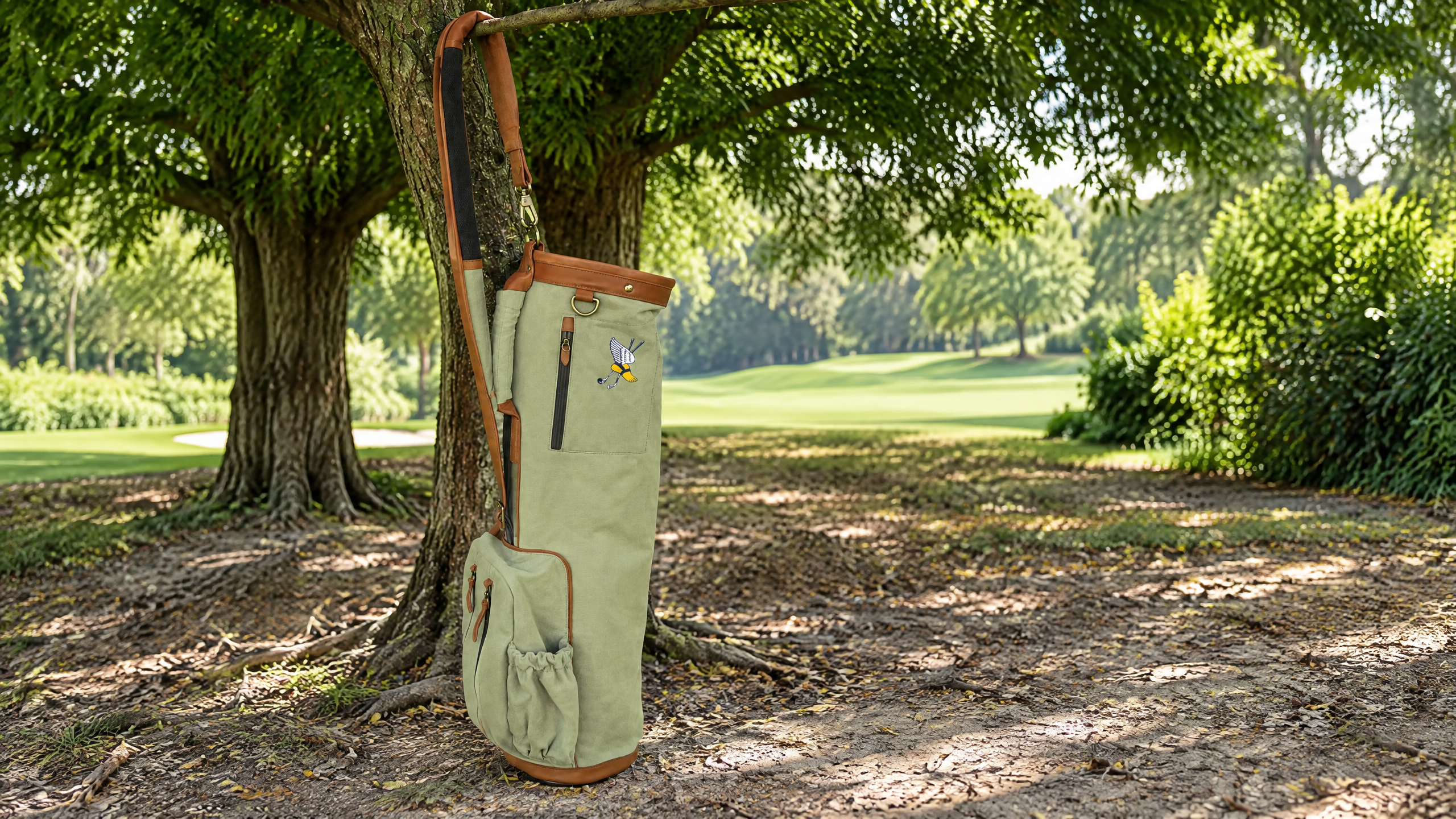 Club-exclusive olive canvas Sunday golf bag with leather trim and embroidered winged-foot crest beside a mature tree on a golf course